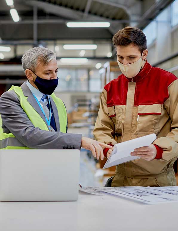 Two workers with face masks discussing items on a notepad in a manufacturing facility